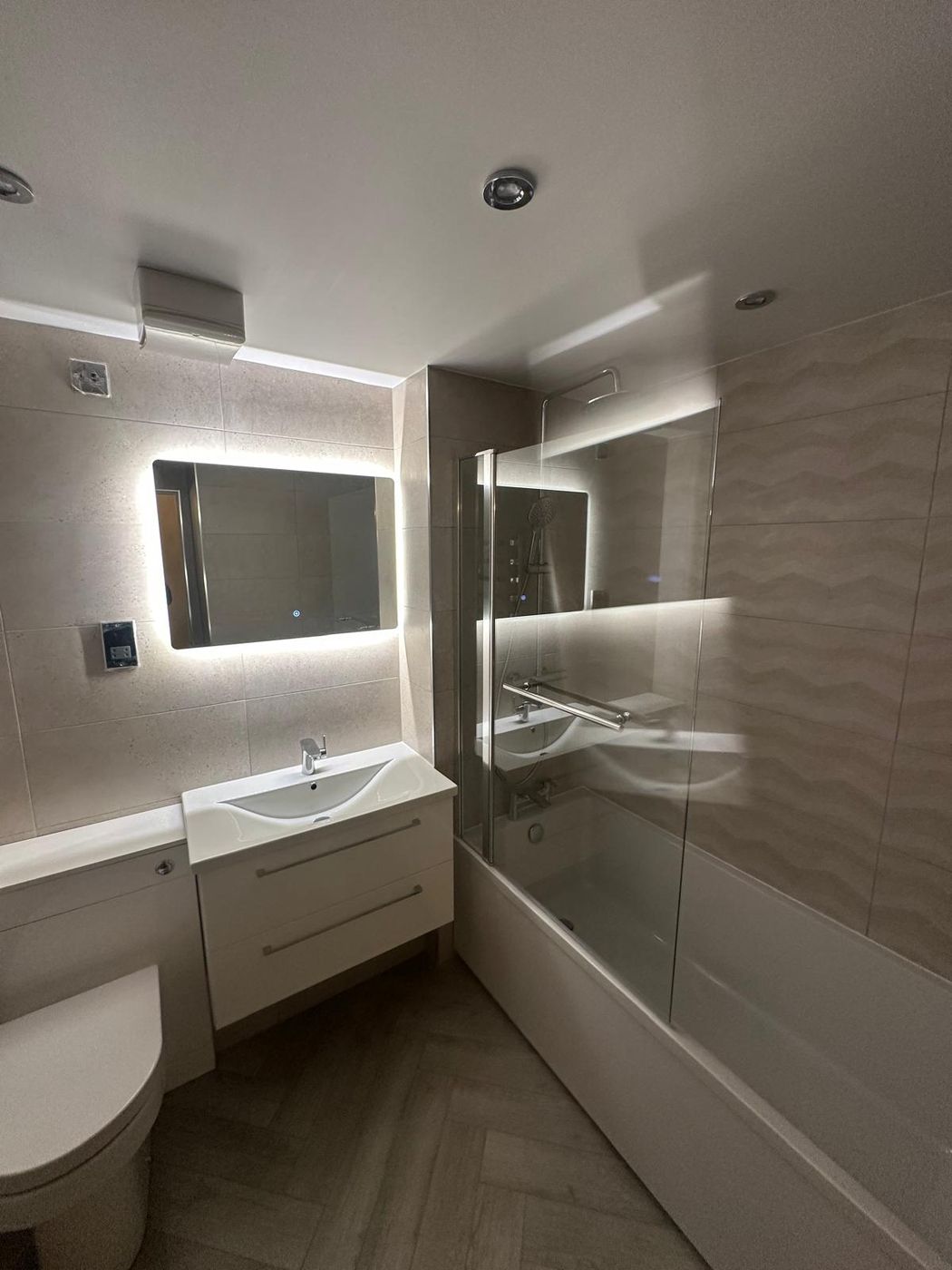 Completed bathroom with backlit illuminated mirror, stone-effect tiling and a bath screen